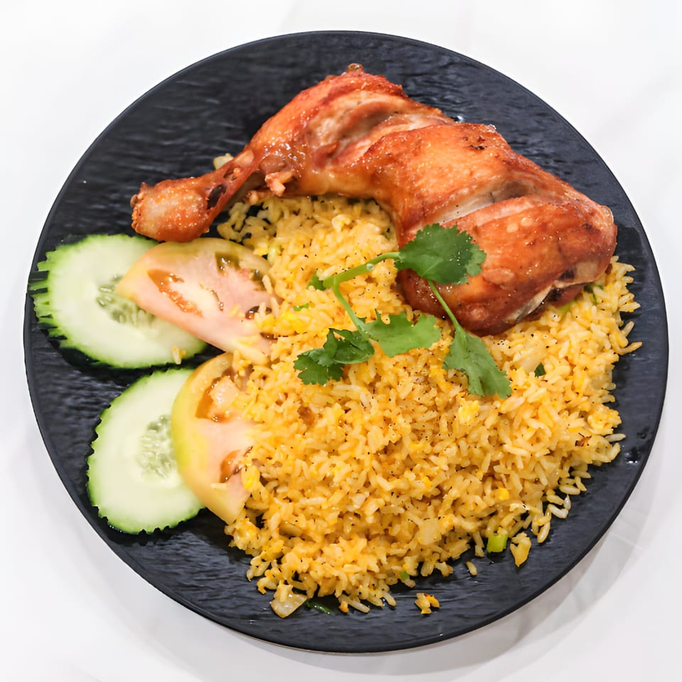Crispy fried chicken leg quarter with yellow fried rice, cucumber, tomato, and cilantro on a black plate.