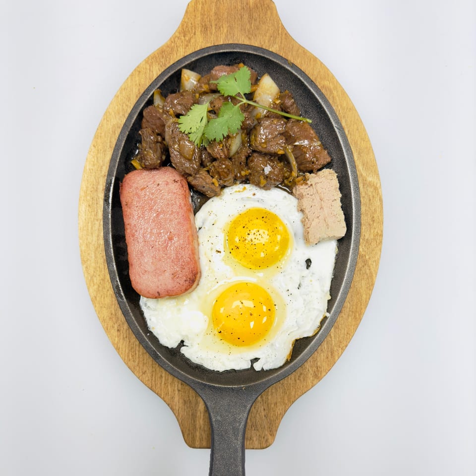 Vietnamese sizzling plate with beef, two fried eggs, spam, and pate, garnished with cilantro.